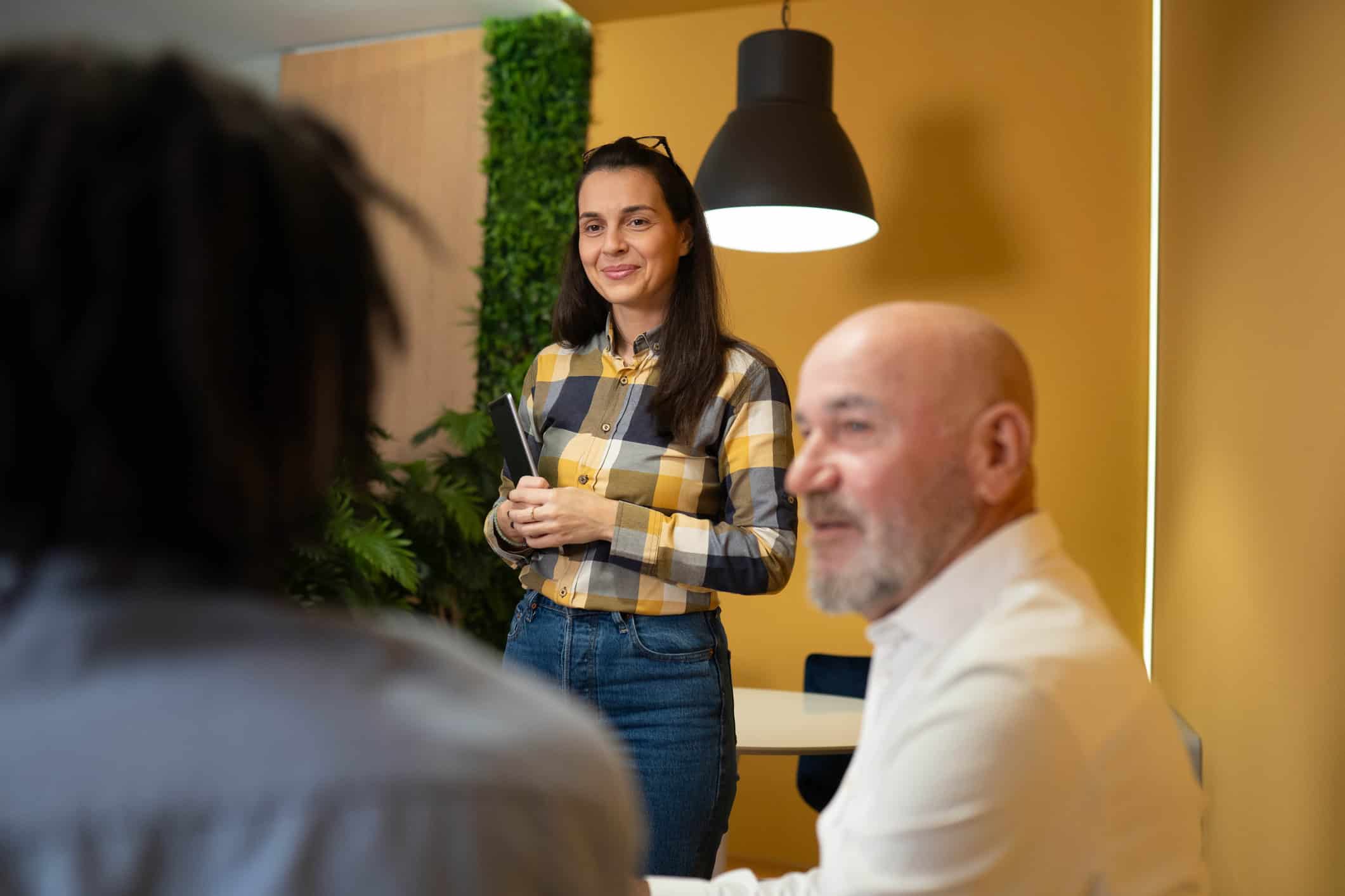 Vrouw in geruite blouse staat ontspannen en zelfverzekerd te glimlachen tijdens een meeting.